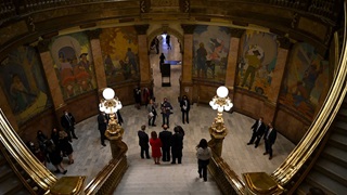 A man in a suit is flanked by colleagues as they pose for a photo at the bottom of a grand staircase. There are colorful paintings of people on the walls in the hall, and other people clad in business outfits watching the photoshoot.