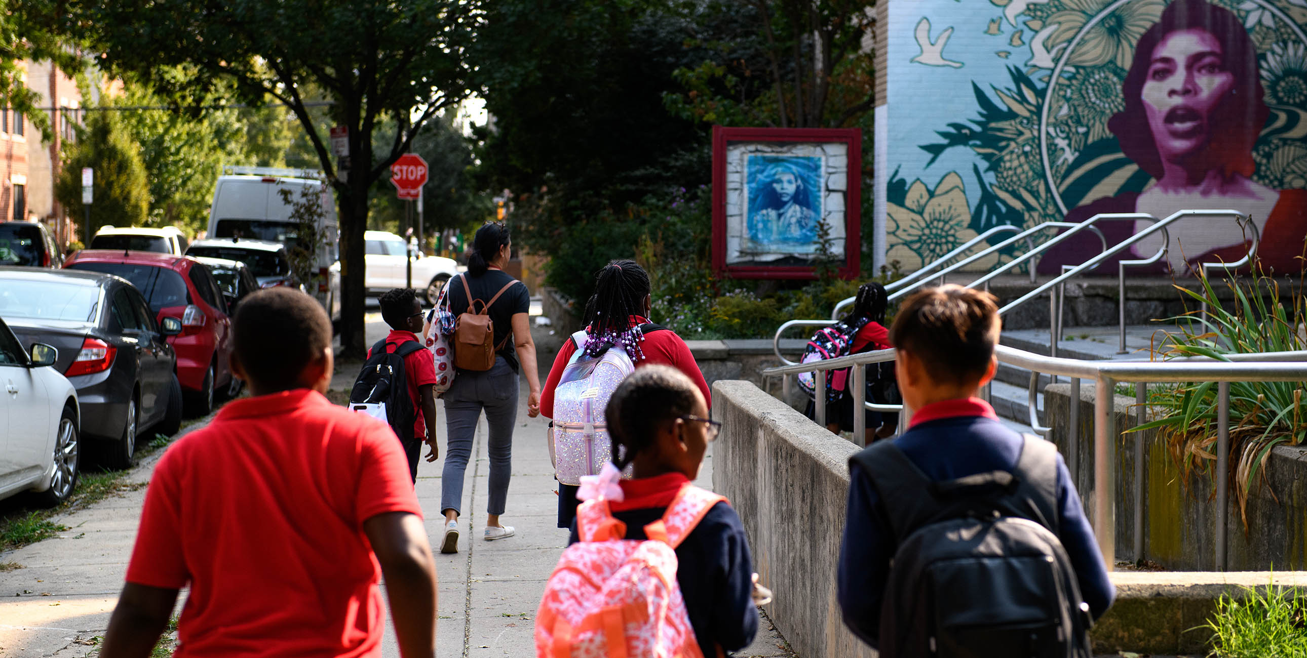 Several children, some wearing backpacks, walk along a suburban sidewalk.