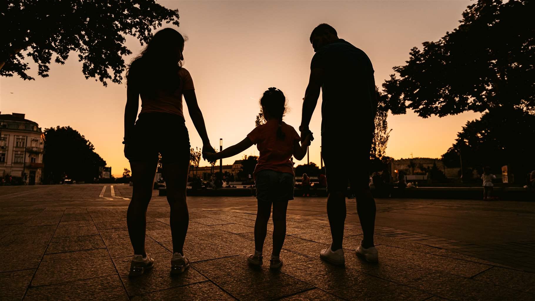 A child holds the hands of two adults, one on either side, as the three stand outside in silhouette against a setting sun.