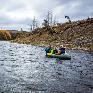 A person paddles an inflatable kayak loaded with camping gear down a river between gently rising banks. The sky above is foreboding, with thick clouds. 