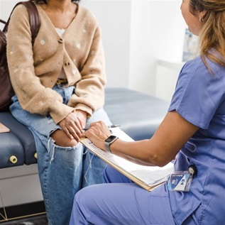 A teenage girl wearing a backpack, tan sweater, and jeans is seen from the neck down sitting in front of a female doctor holding a medical chart with the doctor’s back to the camera.