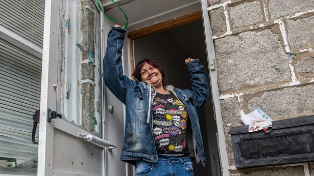 A smiling woman—wearing ripped blue jeans, a black graphic shirt, and a jean jacket—raises her arms and holds up a key while standing in the doorway of a stone building.