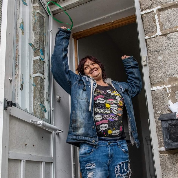 A smiling woman—wearing ripped blue jeans, a black graphic shirt, and a jean jacket—raises her arms and holds up a key while standing in the doorway of a stone building.