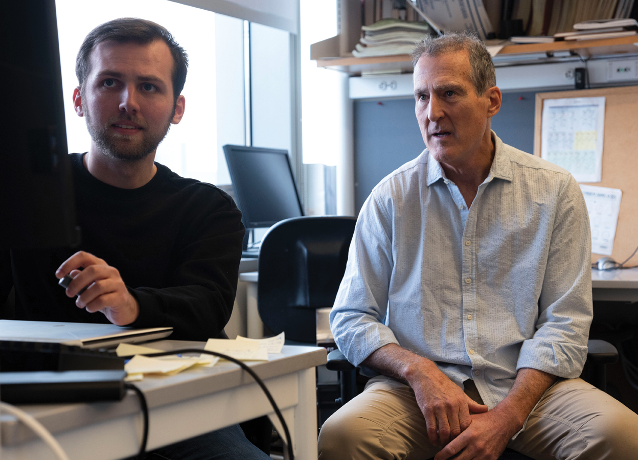 Two people in an office face the camera, consulting a computer monitor barely visible at left.